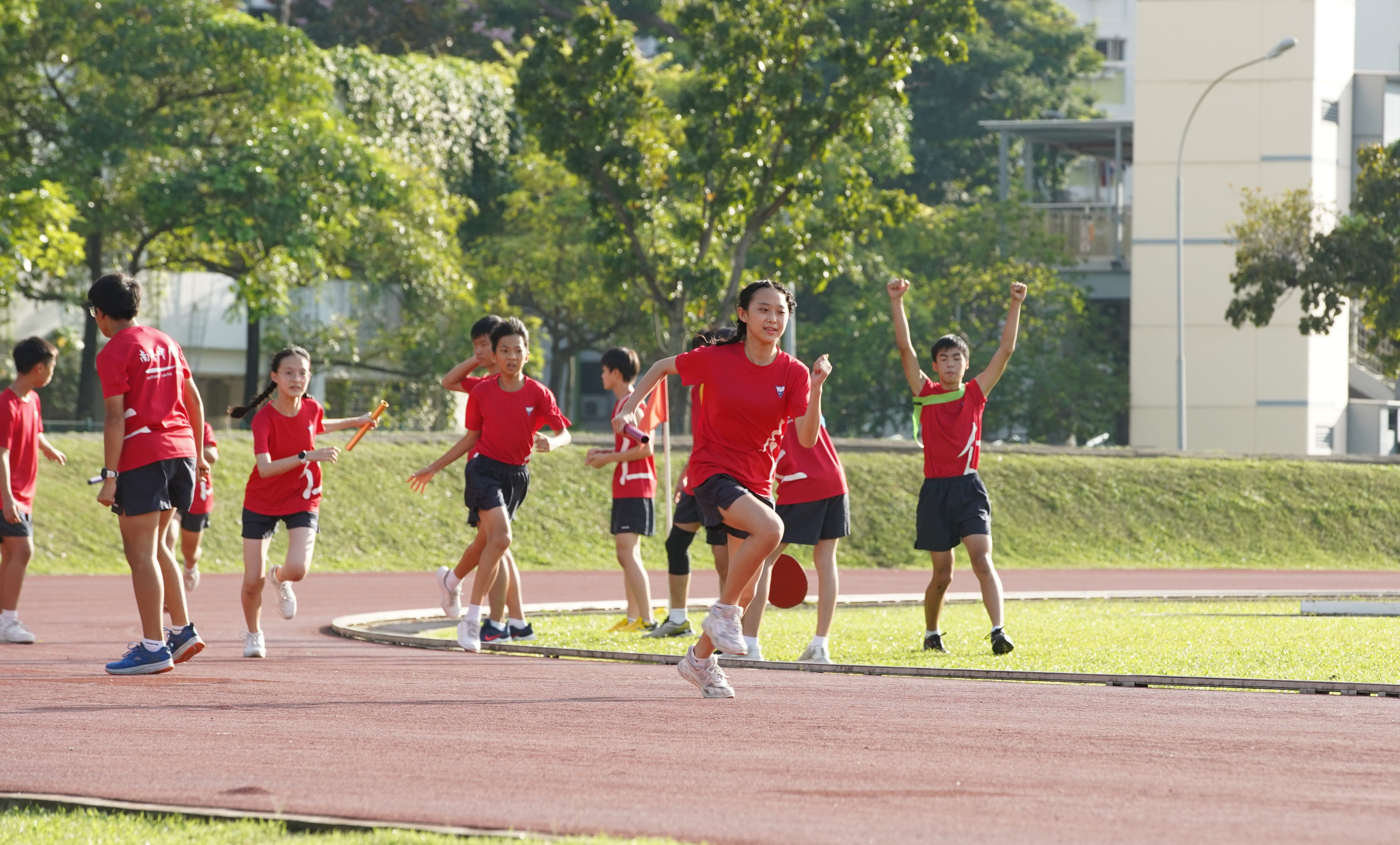 Students in red shirts run a relay race on an outdoor track, with one holding a baton and another cheering.